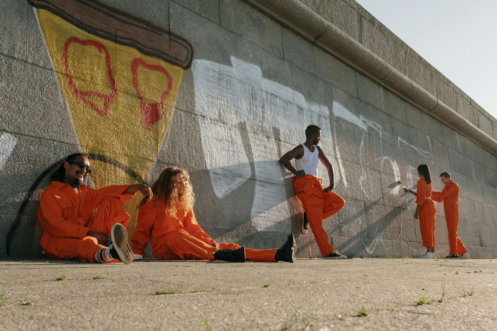 Five people in orange coveralls painting a wall outdoors, showcasing teamwork and diversity.